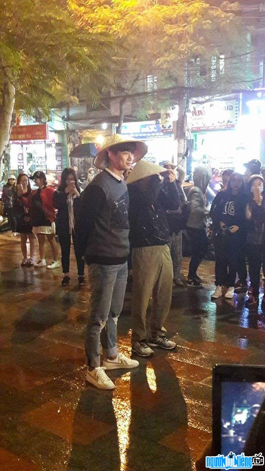 Photo Male singer Ok Taecyeon wearing a hat standing in the rain while filming in Vietnam